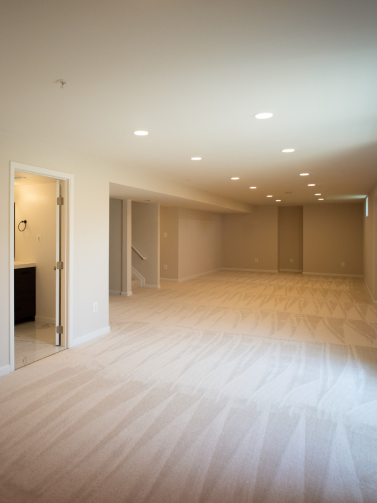A finished basement remodel project featuring wall to wall cream carpeting, recessed lighting, and neutral cream walls.